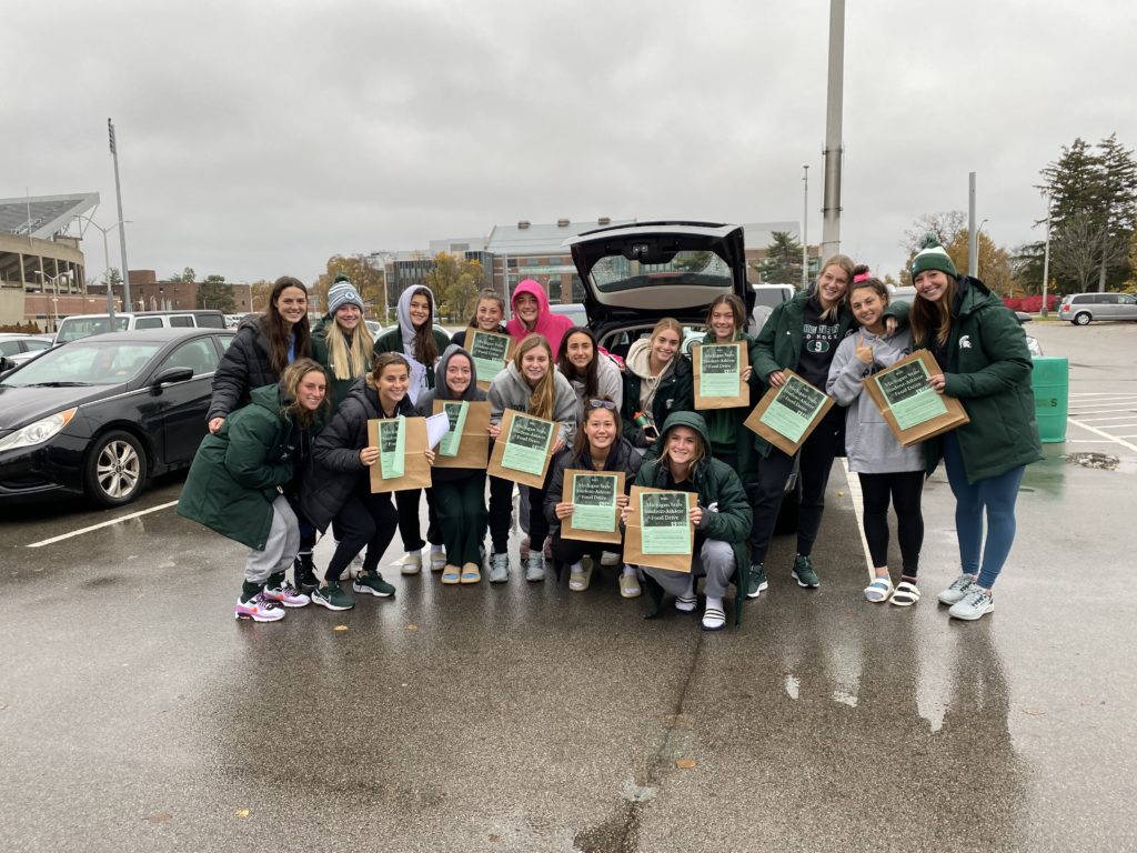 Group of MSU student-athletes pose together in a parking lot, smiling and holding up their food donation bags.