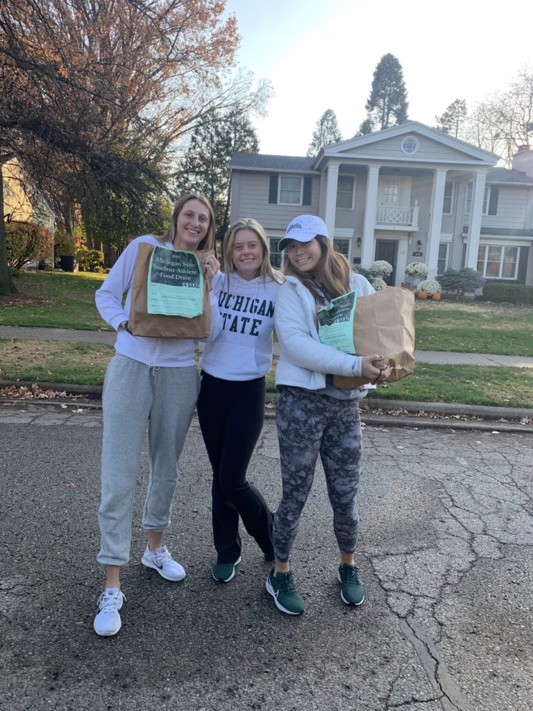 Three MSU student-athletes stand together smiling, holding their food donation bags.