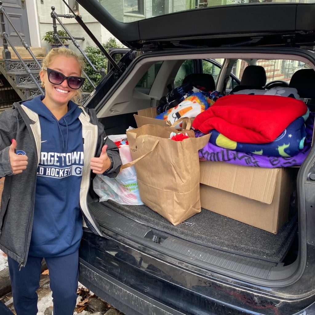 Georgetown field hockey player is smiling and giving thumbs up, posing in front of a car trunk that's filled with blankets the tams made