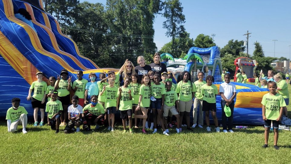 LSU student-athletes pose with about 20 elementary kids all wearing green shirts. They are standing on a field with an inflatable slide behind them.