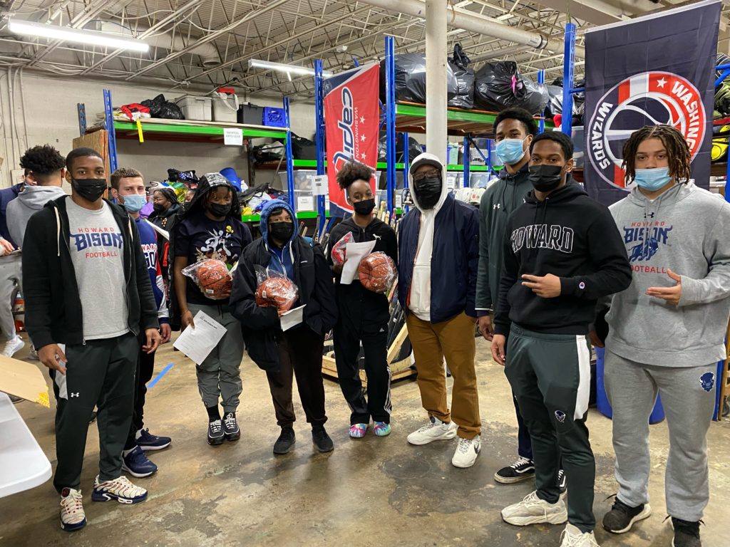 Four Howard football players stand in a warehouse type facility with sports equipment behind them. They are posing with 3 young students from Girls Global Academy.