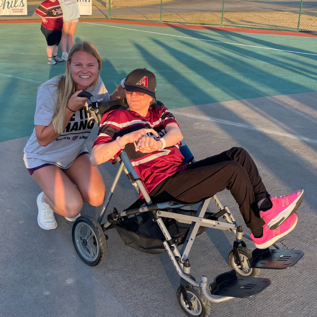 An LSU beach volleyball player squats behind a Miracle League athlete who is in a wheelchair and smiling. They are on a turf baseball field.