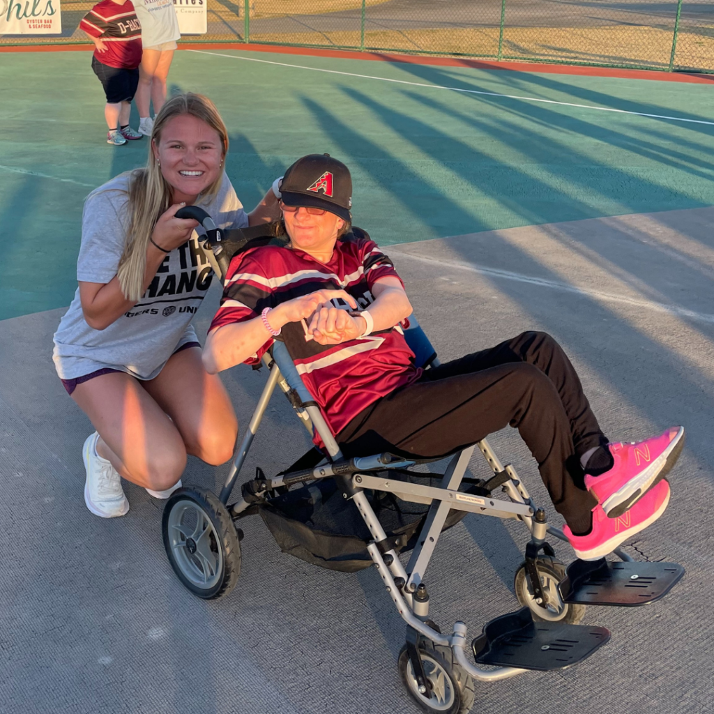 An LSU beach volleyball player squats behind a Miracle League athlete who is in a wheelchair and smiling. They are on a turf baseball field. 