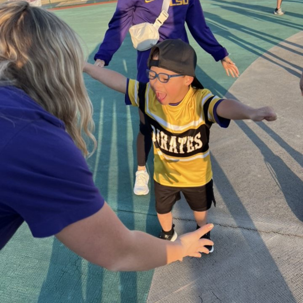 A young Miracle League athlete holds his arms open to hug an LSU student-athlete. He is smiling and they are outside on a baseball field.