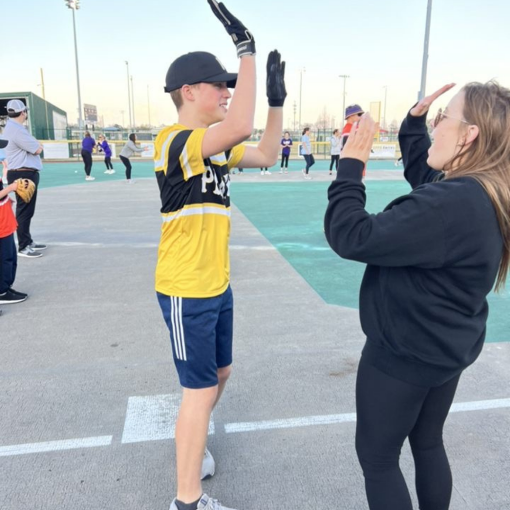 A Miracle League athlete high fives a volunteer. They are standing on third base around a baseball diamond.