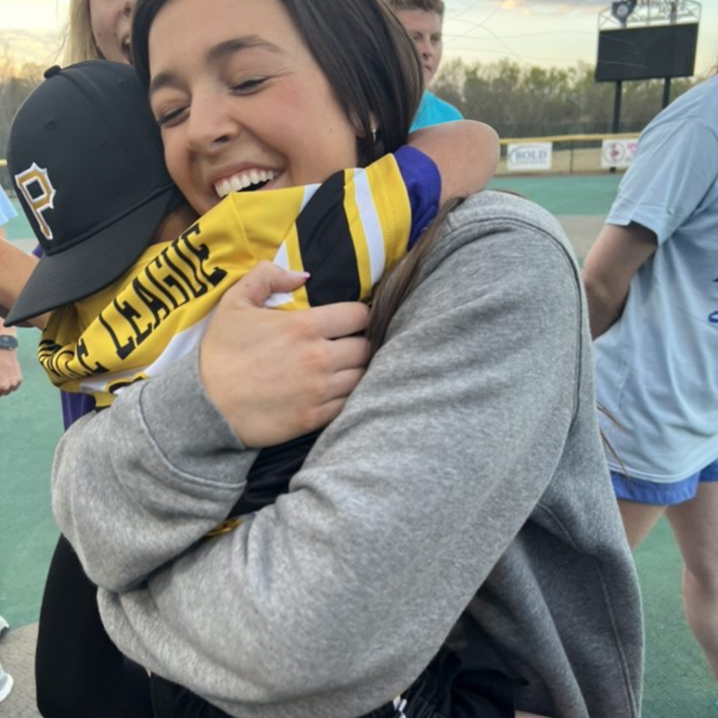 An LSU athlete is smiling with her eyes closed, hugging a young Miracle League athlete.
