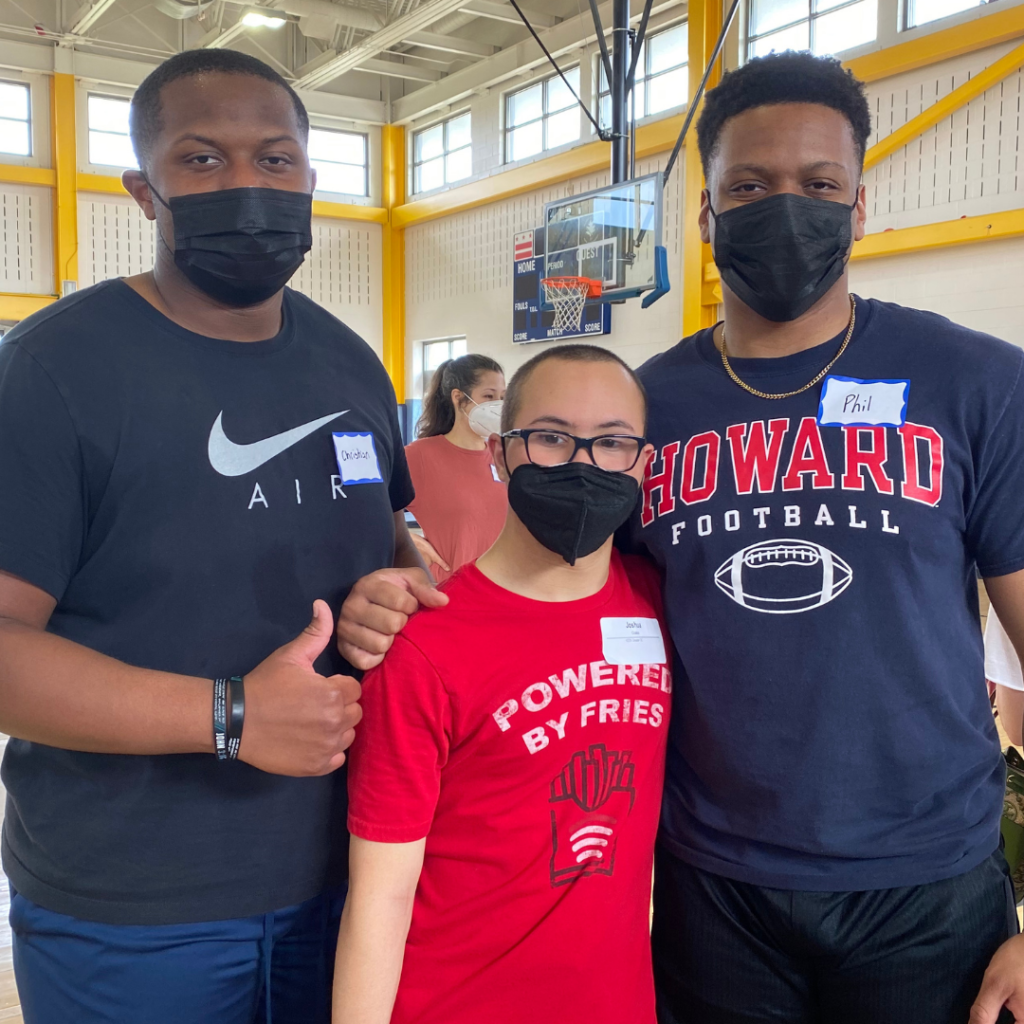 Two Howard football players standing inside a gymnasium posing with a KEEN athlete. A basketball hoop is in the background.