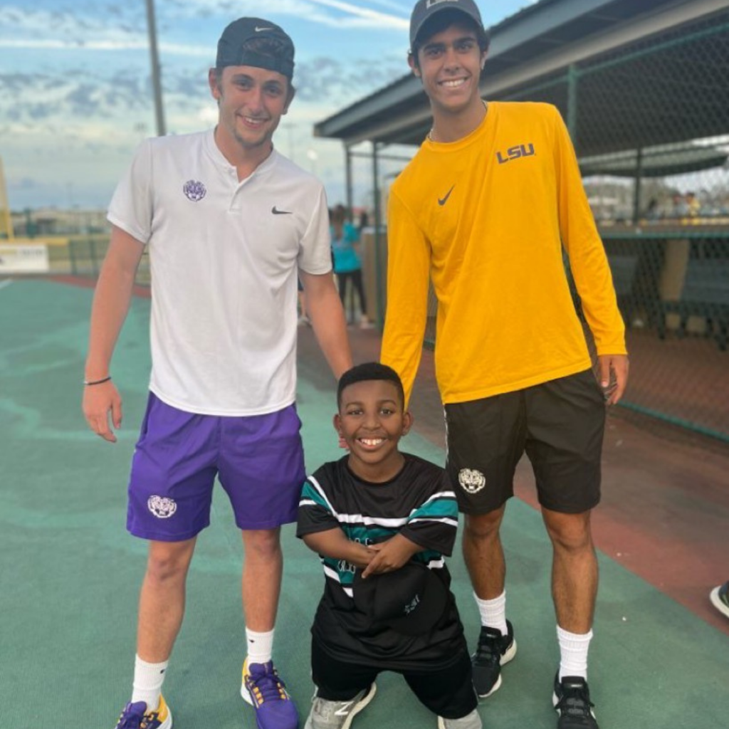 Two LSU tennis players pose with a Miracle League athlete outside on a baseball field. They are all smiling.
