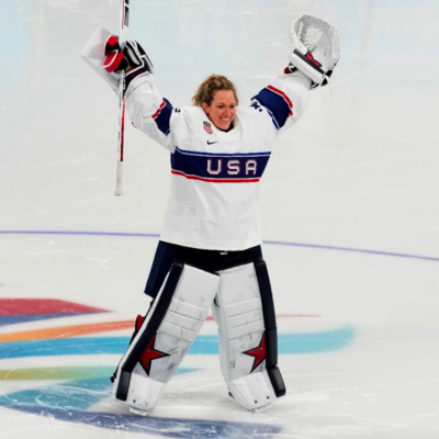 Alex Cavallini stands on an ice rink with her hands in the air, helmet off and smiling.