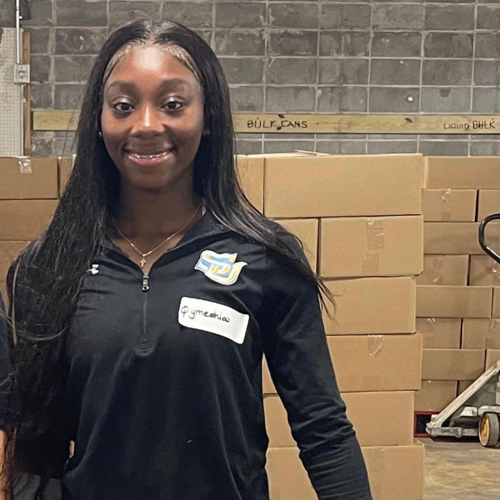 Qymeshia Mayhew smiling in a warehouse-like facility with boxes behind her.