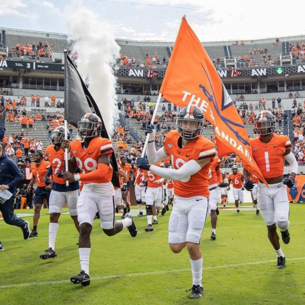 UVA football players hold UVA flags as they run across the football field