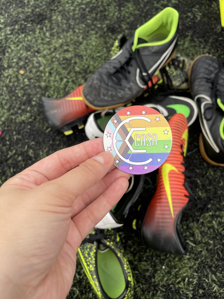 a hand holding a rainbow pride pin over a pile of soccer gear like cleats and shin guards