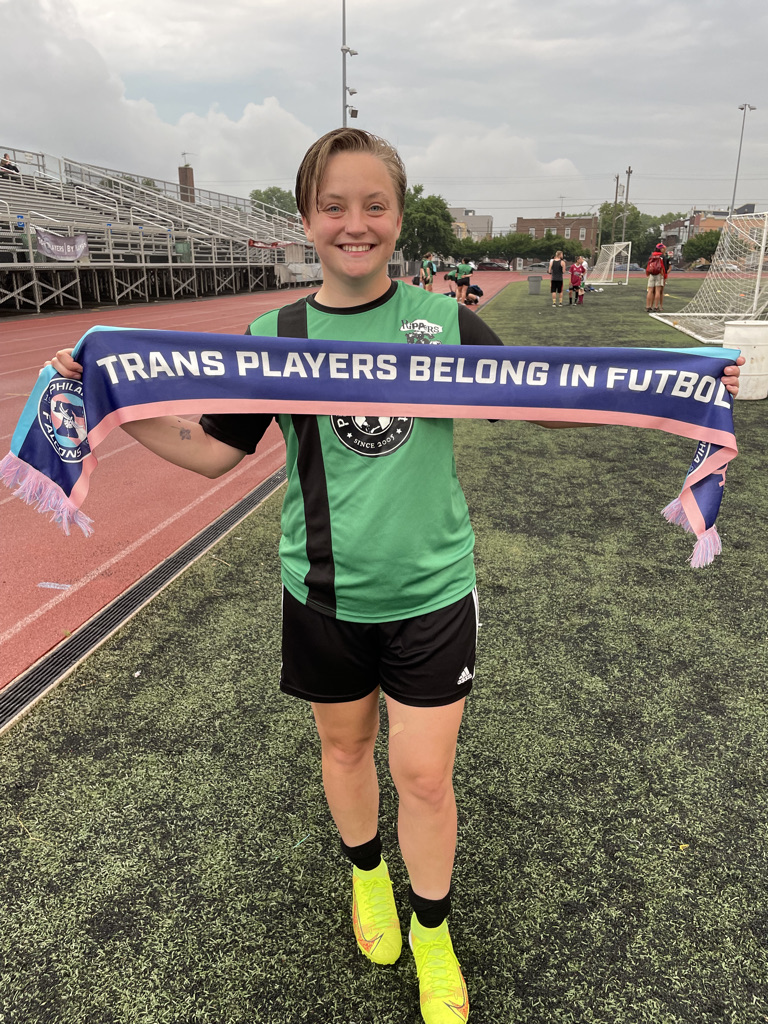 a soccer player holds up a soccer scarf that reads "trans players belong in futbol"