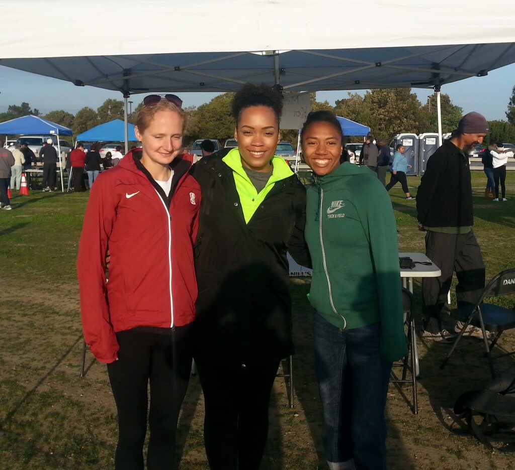 Three women, including Kori, stand on a field in front of an outdoor tent. They are all wearing athletic clothing and smiling at the camera. 