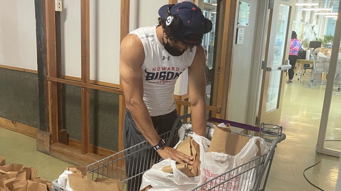 A Howard University football player stands in a hallway while putting items in a grocery bag.