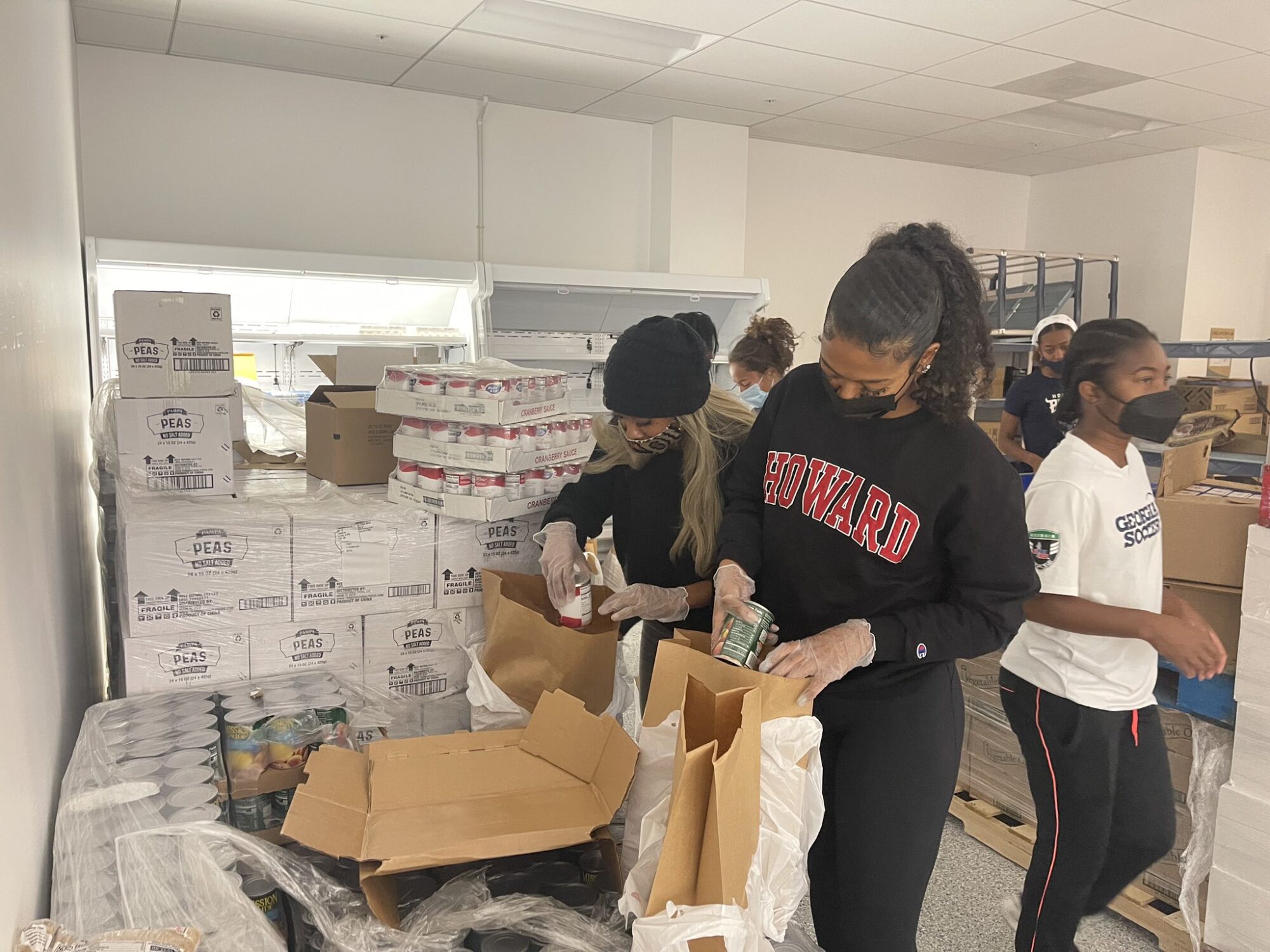 A Howard University student-athlete packs a grocery bag at the Bread for the City facility. Other student athletes stand behind her and there are boxes stacked on the ground.