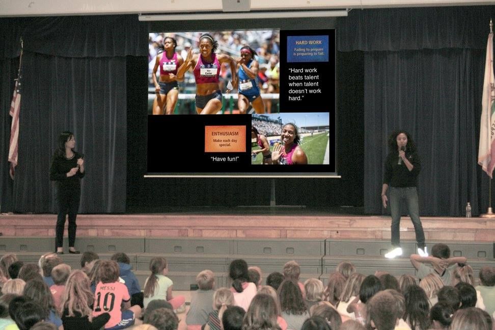 Kori stands on stage holding a microphone in front of a group of children. She is presenting a slide that has photos of herself on it and two quotes. There is another woman standing to the side. 