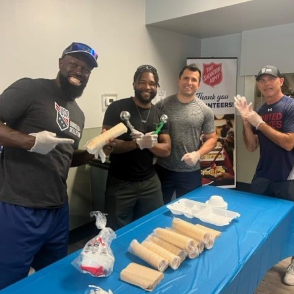 Four members of USABS smile while setting up an ice cream station at the Salvation Army Center of Hope Shelter event. One member is holding up ice cream cones and another is holding up two ice cream scoops.