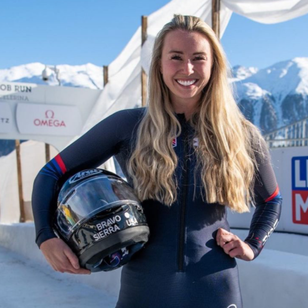 Photo of Riley Tejeck smiling on Bobsled track with helmet under her right arm and left arm on hip.