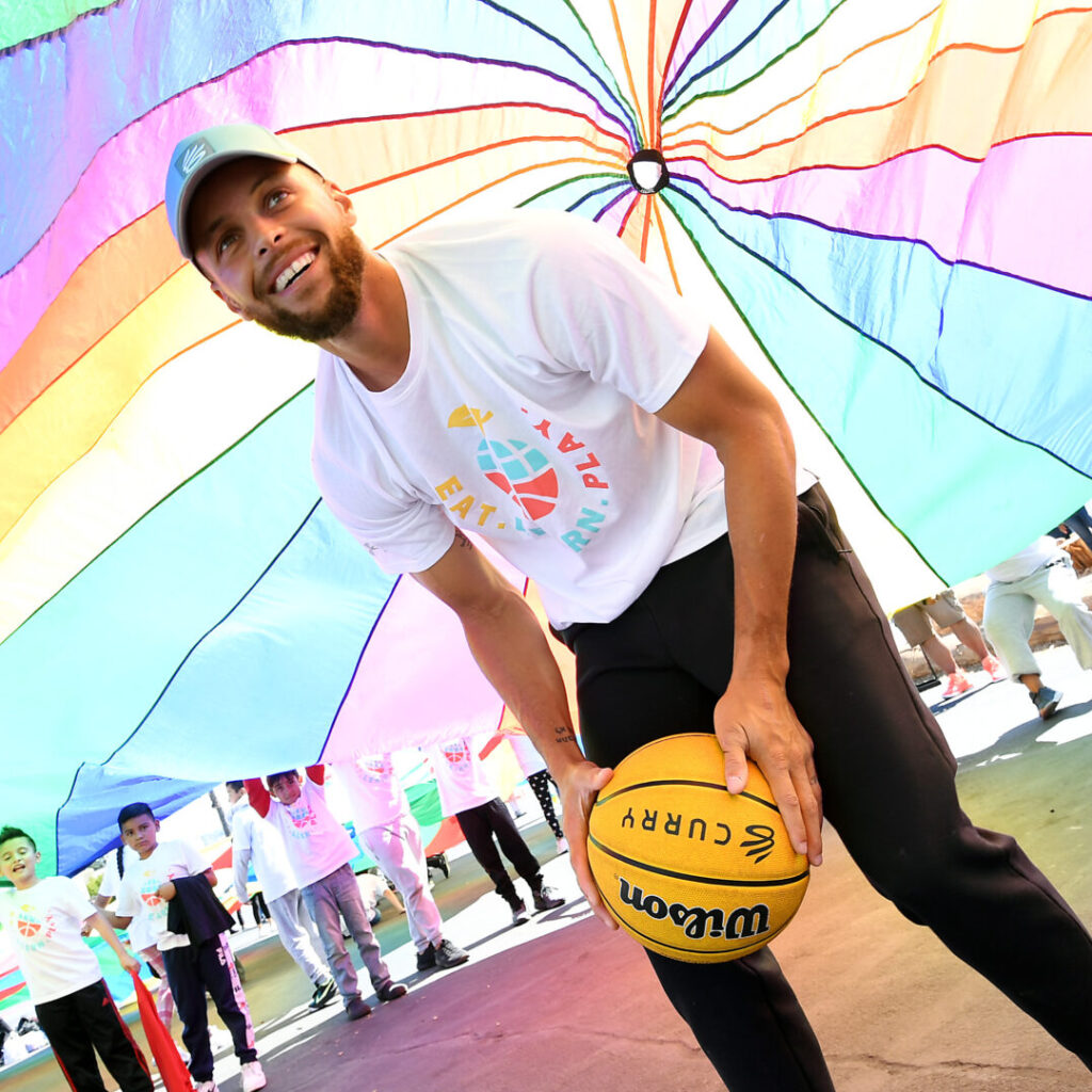 Stephen Curry underneath a rainbow parachute that is being held up by young kids, who can be seen in the background. Stephen is holding a Curry Brand basketball, an Eat. Learn. Play. shirt and a hat. He is smiling up at the camera.
