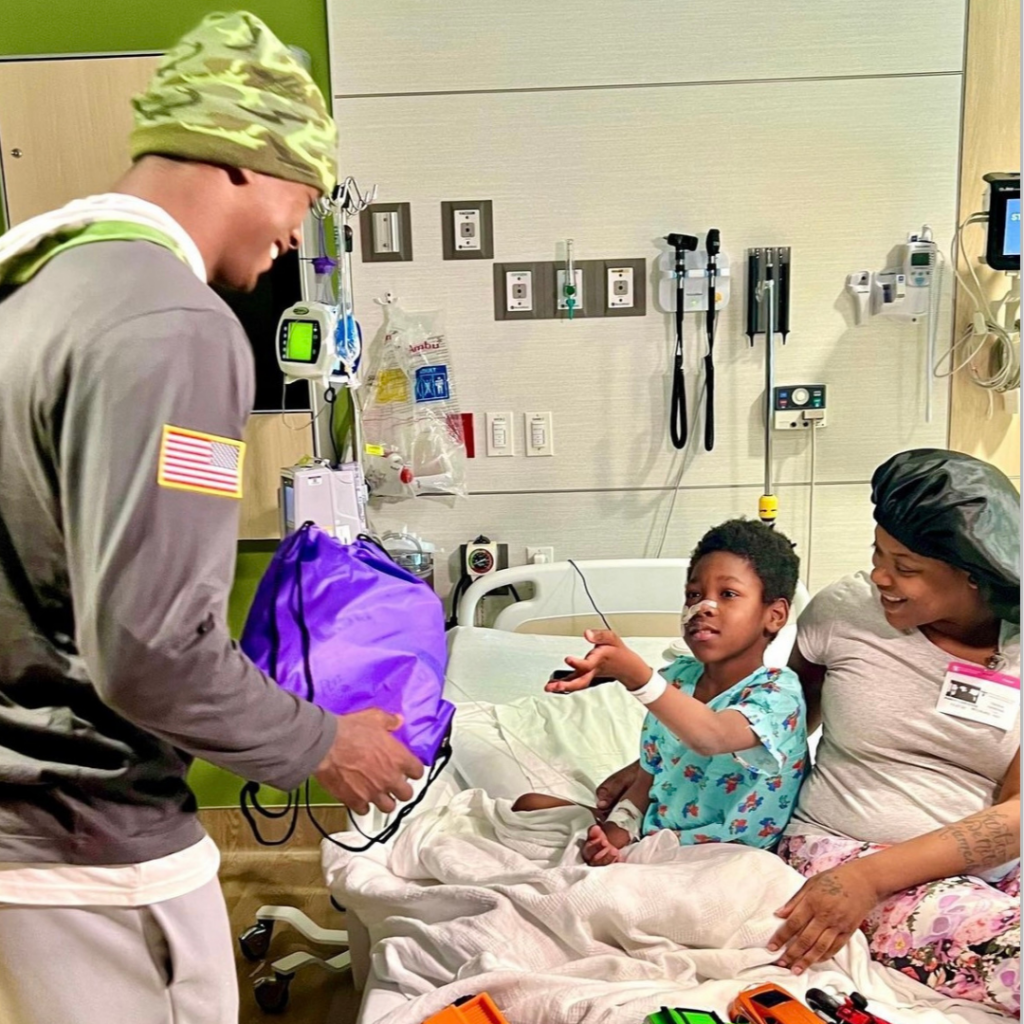 An LSU football member is smiling as he hands a swag bag to a young patient in the hospital.