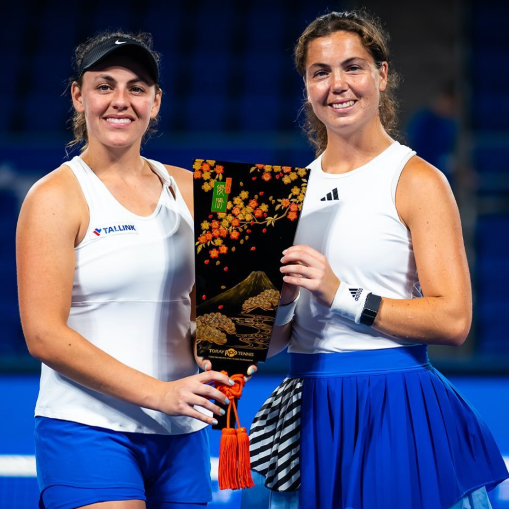 Ingrid Neal poses with her doubles partner Ulrikke Eikeri as they hold up their first place trophy between them. Both girls in white tank tops with blue bottoms.